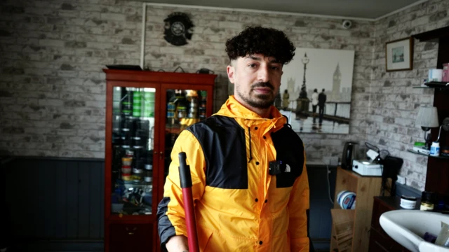 Ahmet Dagelen is wearing a yellow and black rain coat, standing inside his barber shop. He has a mop in one hand.