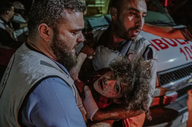 Two members of the Palestinian Red Crescent carry an injured child into hospital in the city of Khan Yunis. The child has long curly hair and is wearing a dazed expression. Her face is covered in dust and blood.