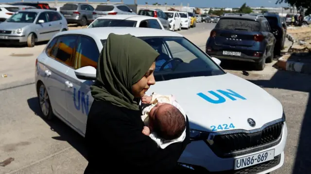 A woman carries a baby as families of staff of international organisations carry their belongings as they shelter at a United Nations center after UNRWA