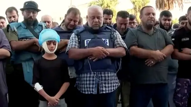 In the middle stands the journalist with his head bowed as he attends his family's funerals. Others stand around him with heads down