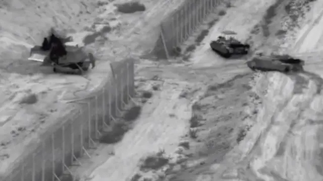 A grainy black and white image of three tanks near a broken security fence