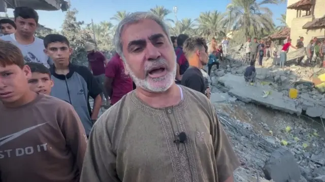 A man speaking to a camera in front of rubble