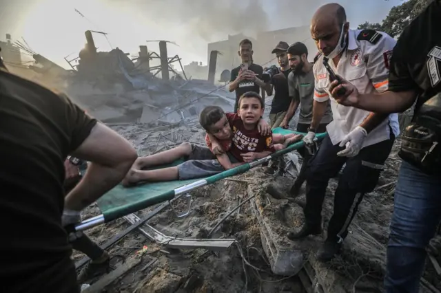 Two boys on a stretcher surrounded by rubble. Men surround them