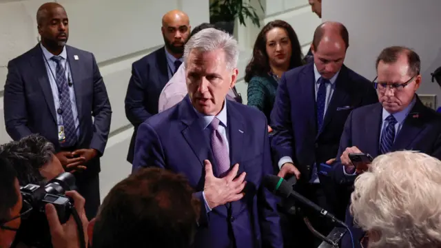 McCarthy in blue suit jacket with purple tie in front of micropohones and reporters