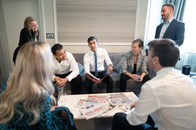 Rishi Sunak sits on a sofa surrounded by staff, in front of a coffee table with newspapers strewn across it