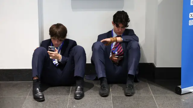 Two men in suits sit on the floor looking at their phones