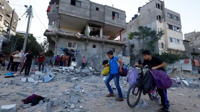 Two young Palestinians walk with bags on ground strewn with debris in front of damaged buildings in Gaza