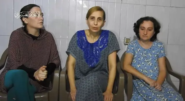 Three hostages sit on plastic chairs in front of a white tiled wall