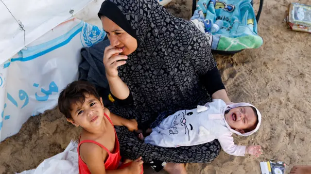 A woman sits with children at a tent camp at a UN-run centre in Khan Younis. She is holding an infant who is crying and a young child is crouched next to her looking up at the camera