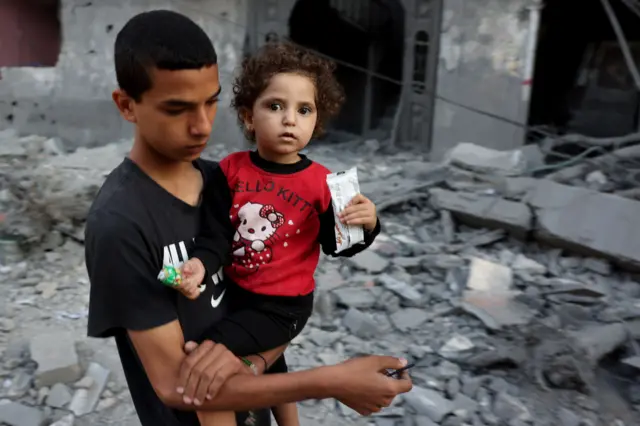 A Palestinian boy carries another young child past destroyed buildings