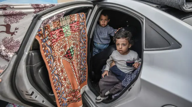 Two young Palestinian children shelter in a car with carpets and rugs hung over the windows to create a make-shift shelter