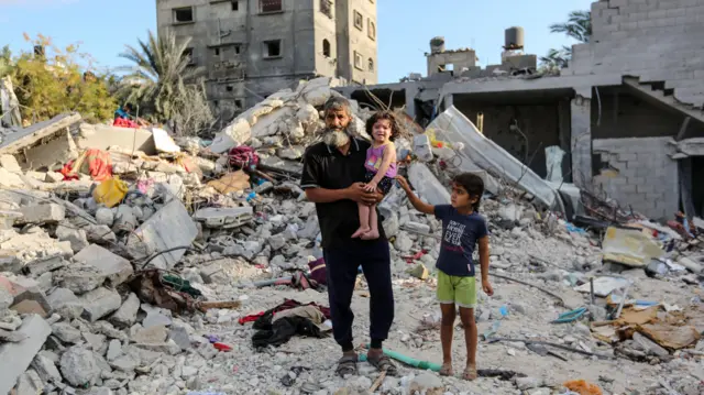 A man stands with a young child on his hip and another standing by his side in Khan Younis. They are standing in a site of rubble where buildings have been bombarded