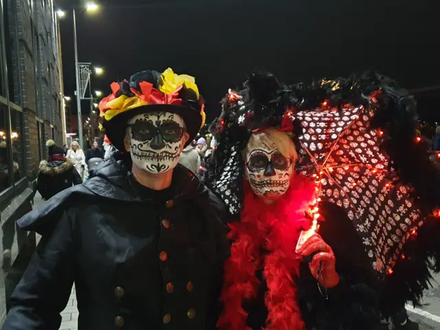 Shows a couple with Day of the Dead masks, with the man on the left wearing a hat and cape and the woman on the right an umbrella