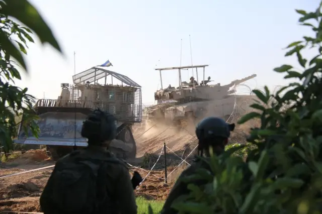 An IDF tank and bulldozer driving, two soldiers in combat gear are in the forground