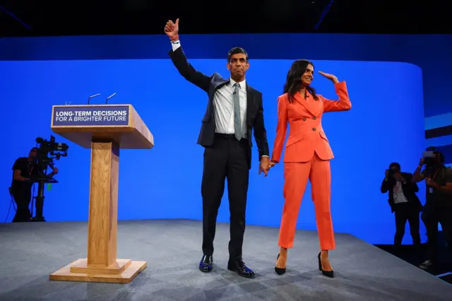 Rishi Sunak gives a thumbs up as he holds hands with his wife on stage at conference