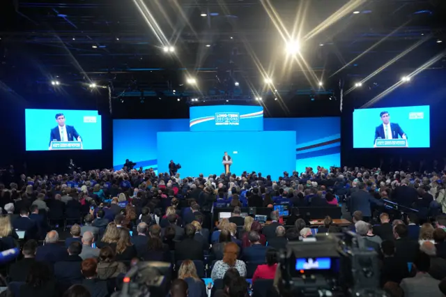 A wide shot of the room, with Sunak on stage and two big screens either side of him