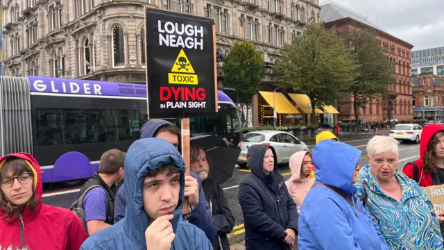 A group of protesters outside Belfast City Hall, holding a placard that says "Lough Neagh TOXIC Dying in Plain Sight"