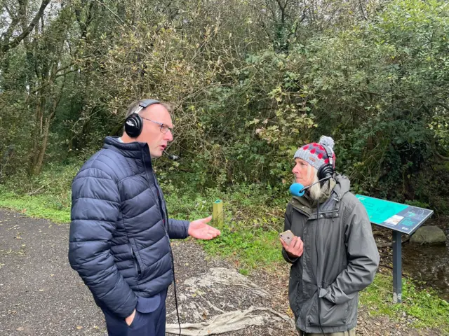 Keith Buchanan and Sarah Brett in conversation, both wearing warm coats and headphones