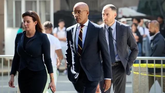 Damian Williams in center, wearing glasses, dark suit, striped tie, between a woman in a blue jacket and man in grey suit.