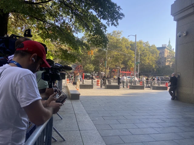 Man with red baseball cap leaning against police fence, cameras on tripods, open square