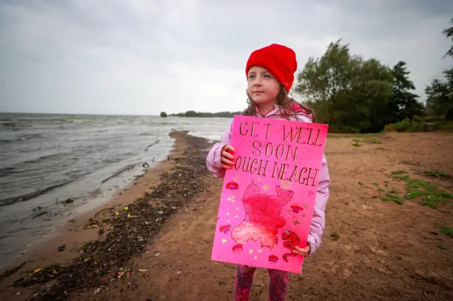 A girl holds a placard that reads "Get well soon Lough Neagh" as she stands on the lake's shoreline