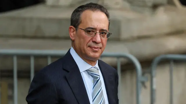 Mark Cohen in front of security fencing, wearing dark suit, blue shirt, striped tie and glasses