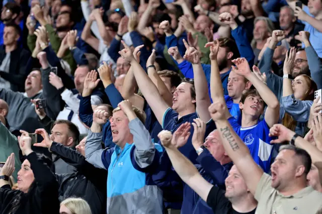 Everton fans raise their arms to clap and cheer their side.