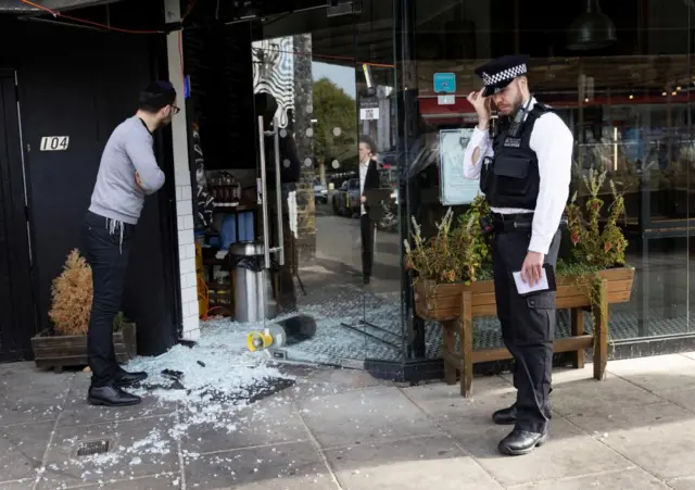 A man and a police officer stand next to a vandalised Kosher restaurant in Golders Green, London