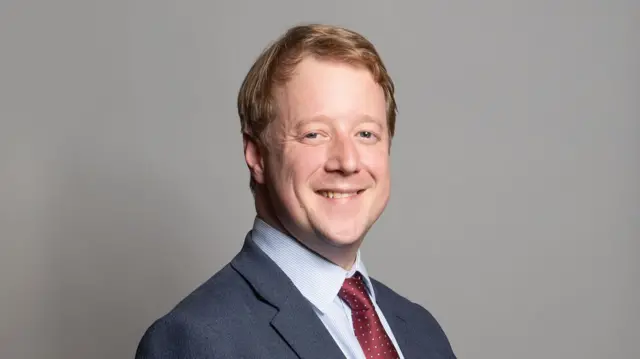 Paul Bristow, official parliament photo, he is wearing a dark suit, light blue shirt and red tie.