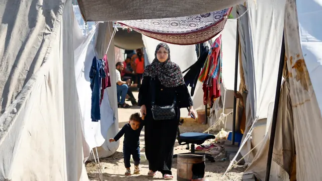 A woman walks with a child as displaced Palestinians, who fled their houses amid Israeli strikes, take shelter at a tent camp at a United Nations-run centre, in Khan Younis, Gaza