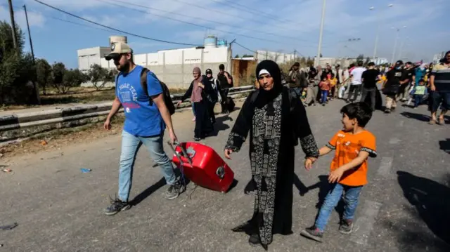 A man drags a suitcase as a woman holds the hand of a child - they walk in front of a crowd down a road