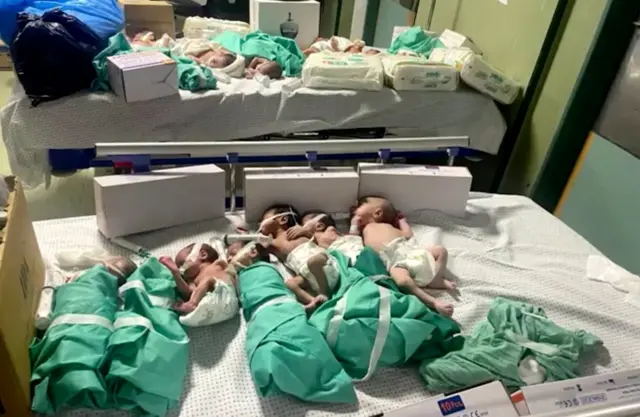 A group of babies, some wrapped in swaddles, some only in nappies, seen lying on hospital beds