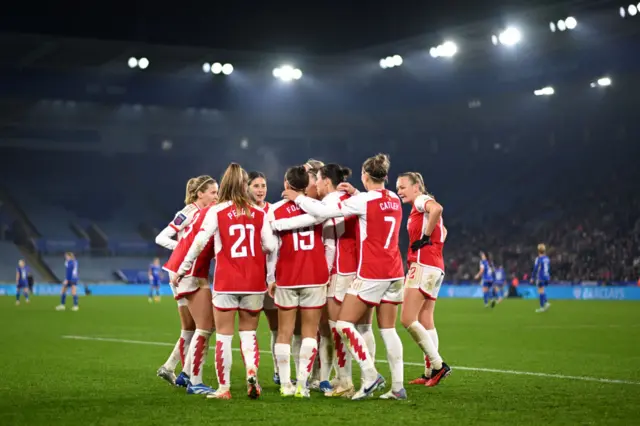 Arsenal players celebrate their goal as a group with the King Power covered in rain in the background.