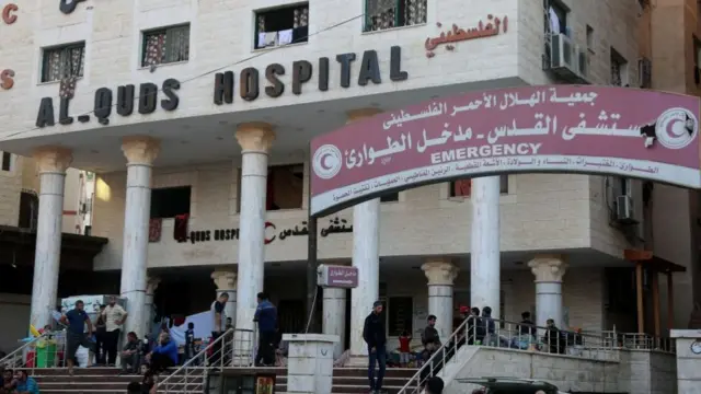 People wait on steps outside a hospital. A banner nearby points to the emergency department