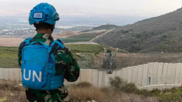 A soldier in a blue UN bullet proof vest looks out over a concrete wall that separates Lebanon and Israel