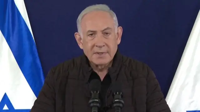 Benjamin Netanyahu speaks into microphones in front of an Israeli flag