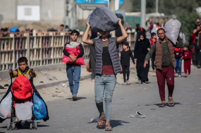 A man carries a bag over his head as he walks down a road. Nearby, a child wheels a pram and others carry various bags