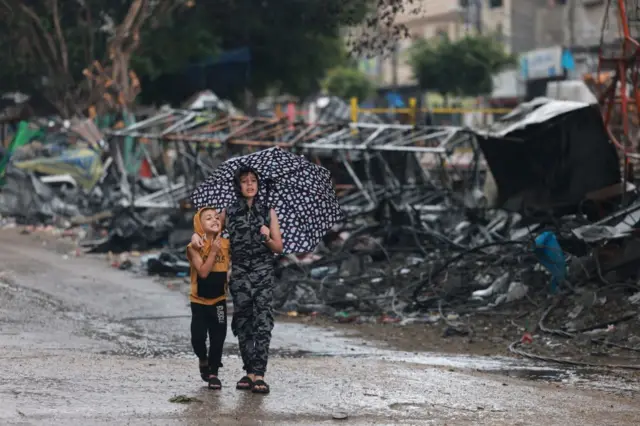 Two Palestinian children walk under an umbrella following rains along a street covered in debris in Rafah in the southern Gaza Strip