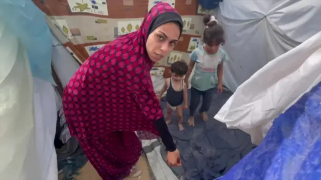 A Palestinian woman gestures to a sheet on the ground of a makeshift tent which two of her young children are standing on