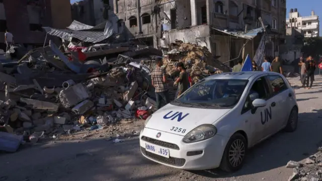 A UN branded car drives past a pile of rubble taller than the car.