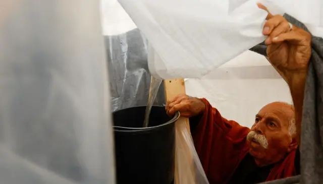 A man pulls the corner of a white tent down, collecting the ensuing stream of brown water into a bucket