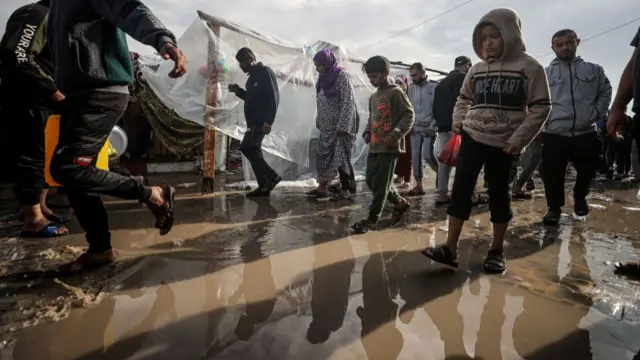 A crowd of people step over a muddy puddle between tents