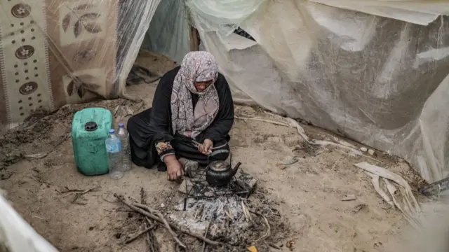 A woman sits on a dirt floor. In front of her, a black teapot is perched on a makeshift camp fire. A filthy jerry can sits beside her