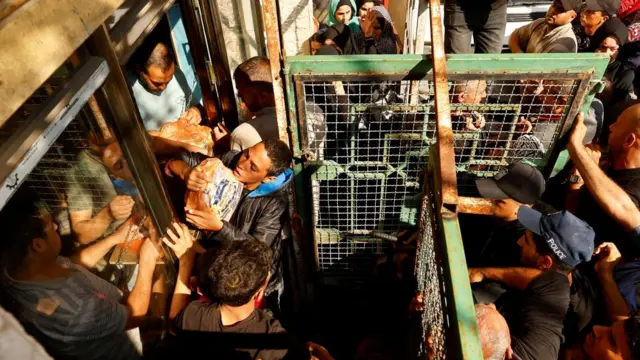 A man is handed a bag of bread through a shop window. A green fence partitions the shop from the crowd