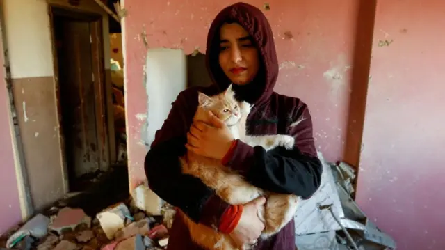 A woman holding a cat in a room full of rubble