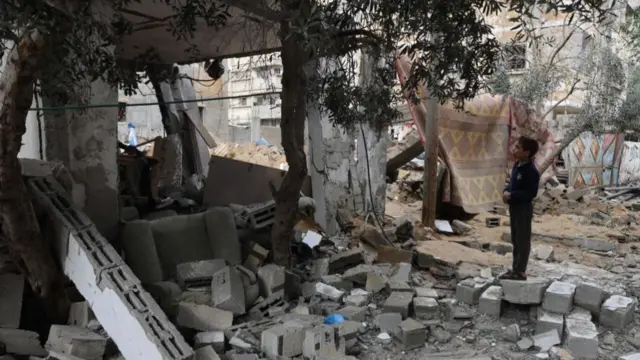 A young child stands on a brick, looking up at the shell of a house. A tree has survived the strike and grows from the rubble