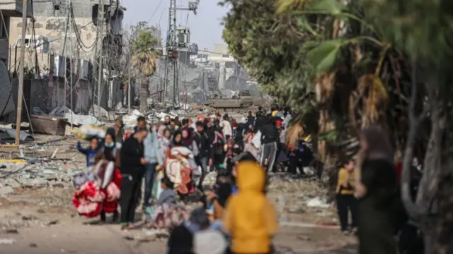 A crowd of people walk past rubble. In the background, in focus, an Israeli tank is parked up the street