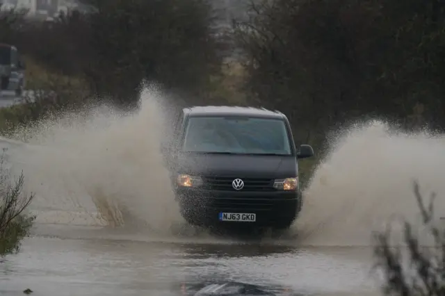 A van drives through flood water on a road and water splashes high on either side