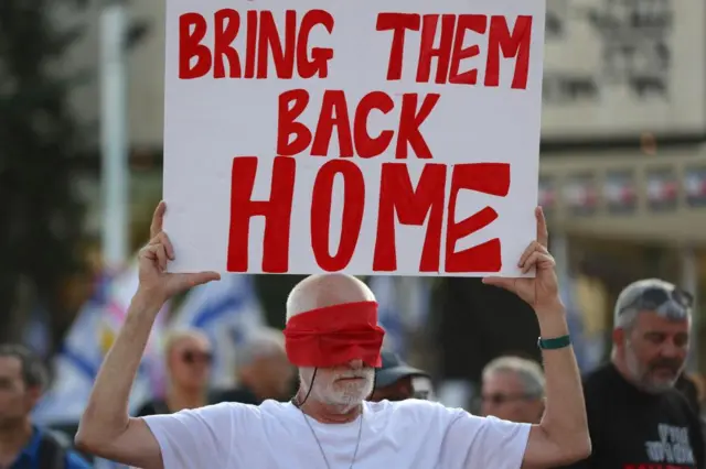 A man with red fabric tied around his eyes holding a sign reading 'bring them back home'.