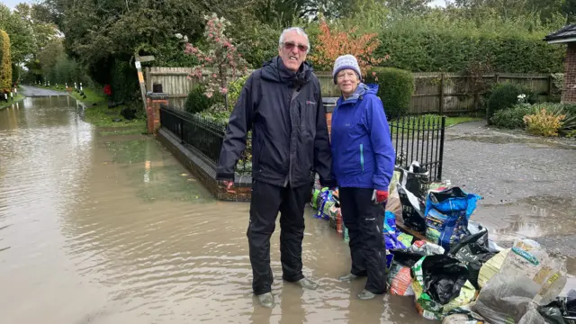 Peter and Hilary Thomas outside their front garden gate which has bags of compost and sand piled up.